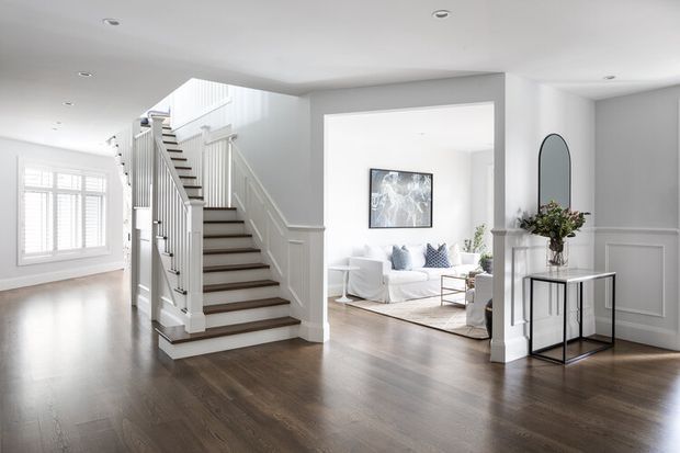 Bright, refined residential entry hall with dark timber flooring, white painted walls and decorative wall panelling. A central staircase with white balustrades and dark treads leads upward, while a large opening reveals an adjoining living room with light furniture and patterned cushions. Plantation shutters, classic trim, a slim console table and arched mirror complete the polished interior.