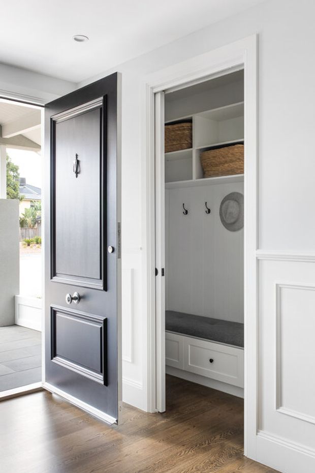 Bright transitional entry area with a dark paneled front door and white trim, opening to a built-in mudroom nook. The mudroom contains a cushioned bench, lower drawers, open shelving with woven baskets, and wall hooks. Timber flooring, white wall panelling, and a glimpse of the exterior create a clean, refined residential entry.