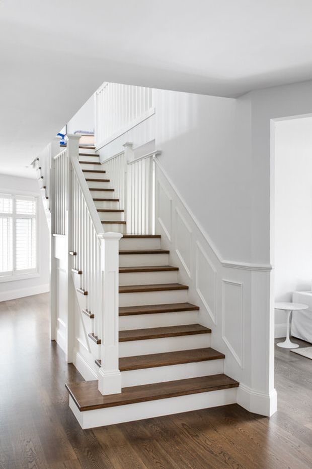 Bright interior view of a residential staircase and landing. The staircase has dark stained timber treads, white painted risers, detailed white balustrades, turned newel posts, and paneled wainscoting. Dark timber flooring continues through the open hall, with a shuttered window on the left and a partially visible sitting area through the opening on the right.
