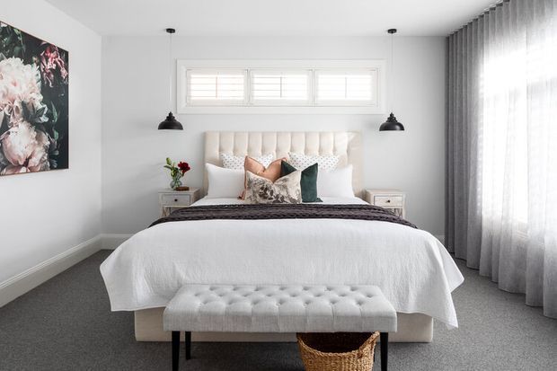 Bright, softly styled bedroom with a large upholstered bed, layered decorative cushions, tufted bench, bedside tables, pendant lights, floral artwork, and full-height sheer curtains. The room has a neutral palette with dark accents and grey carpeting.