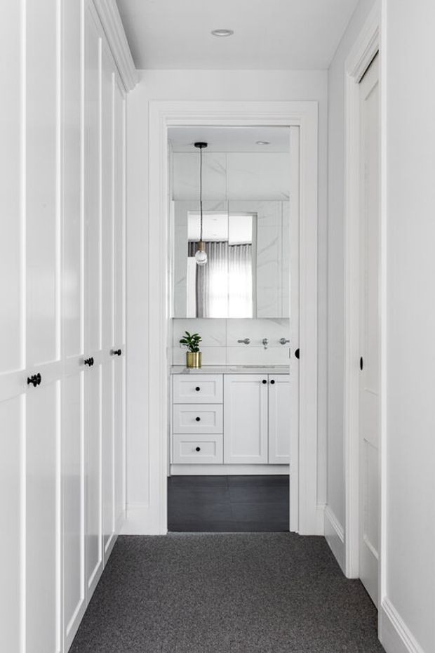 A bright, narrow hallway lined with white built-in panelled cabinetry and white trim, leading directly into a compact white bathroom. The bathroom includes a mirrored wall, white vanity cabinetry, chrome tapware, a small potted plant, dark flooring, and a pendant light.