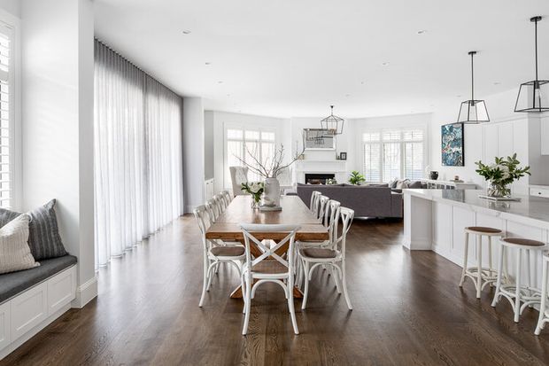 Bright open-plan interior combining a dining area, kitchen and living room. The space features dark timber flooring, white cabinetry and walls, plantation shutters, sheer full-height curtains, a large dining table with white chairs, a grey sofa, and pendant lighting. The styling is refined and coastal-inspired with abundant natural light and a spacious layout.