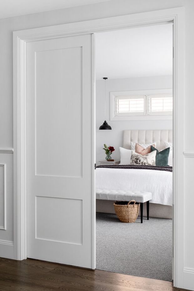View through a white paneled doorway into a bright, softly styled bedroom. The bedroom features a tufted upholstered bed with white bedding and layered teal, blush, and patterned cushions, a grey upholstered bench, a woven basket, grey carpet, a black pendant light, and a high horizontal window with white shutters. The foreground hallway has dark timber flooring and white decorative wall trim.