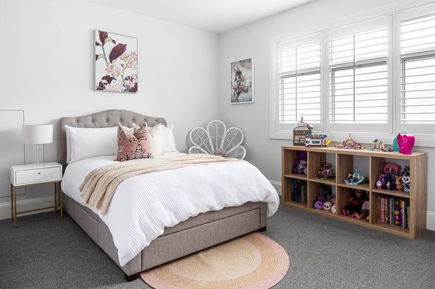 Bright, tidy children's bedroom with a large upholstered bed, white bedding, blush and floral accent cushions, grey carpet, bedside table and lamp, butterfly-shaped decorative chairs, and an open wooden cubby shelf filled with toys and books. White plantation shutters provide abundant natural light, while botanical artwork adds colour.