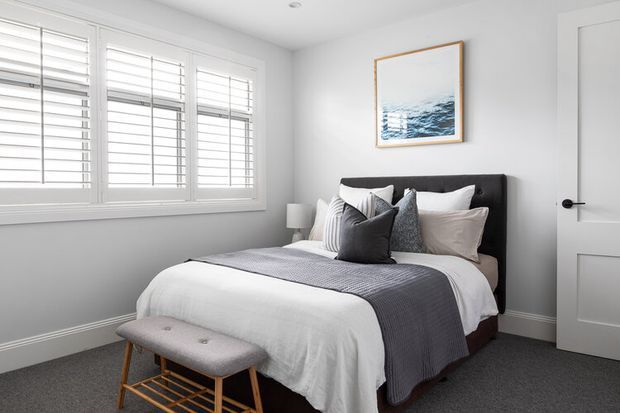 Bright, compact bedroom with a neatly made upholstered bed, layered white and grey bedding, a grey upholstered bench with timber legs, plantation shutters, white painted walls and trim, and a framed coastal artwork above the bed. The room has a calm, refined, contemporary-traditional feel.