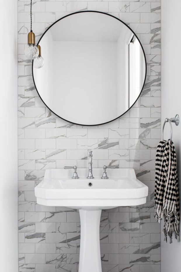 A compact, bright bathroom featuring a white pedestal sink with chrome tapware, marble-look white and grey subway tile walls, a large round black-framed mirror, brass-and-glass pendant lights, and a monochrome patterned towel.