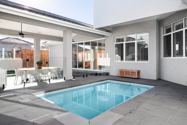 Bright contemporary residential courtyard with a rectangular swimming pool surrounded by large grey paving slabs. The space features white rendered walls, extensive glazed doors and windows, an outdoor dining area under a covered section, ceiling fans, and timber fencing visible beyond the courtyard.