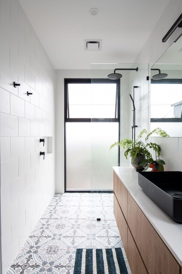 A bright, narrow contemporary bathroom with white rectangular wall tiles, patterned encaustic-style floor tiles, black-framed frosted glazing, and a clear glass walk-in shower. A long timber vanity with a pale benchtop supports a large matte-black vessel basin and black tapware. A large mirror reflects the shower and window, while potted greenery adds a natural touch.