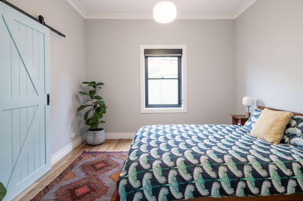 A bright, compact bedroom with a patterned bedspread, a timber barn-style sliding door, a central window with a dark roller blind, a potted plant, bedside lighting, and layered patterned rugs. The room has soft grey walls, white trim, and a simple pendant light, creating a relaxed contemporary look with colourful textile accents.