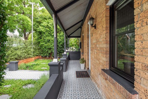 Covered veranda running along a textured brick-clad home, with patterned tiled flooring, dark painted balustrade and posts, timber-lined pitched canopy, black-framed windows, and a landscaped garden beyond. The space has a traditional, sheltered outdoor character with strong geometric detailing.