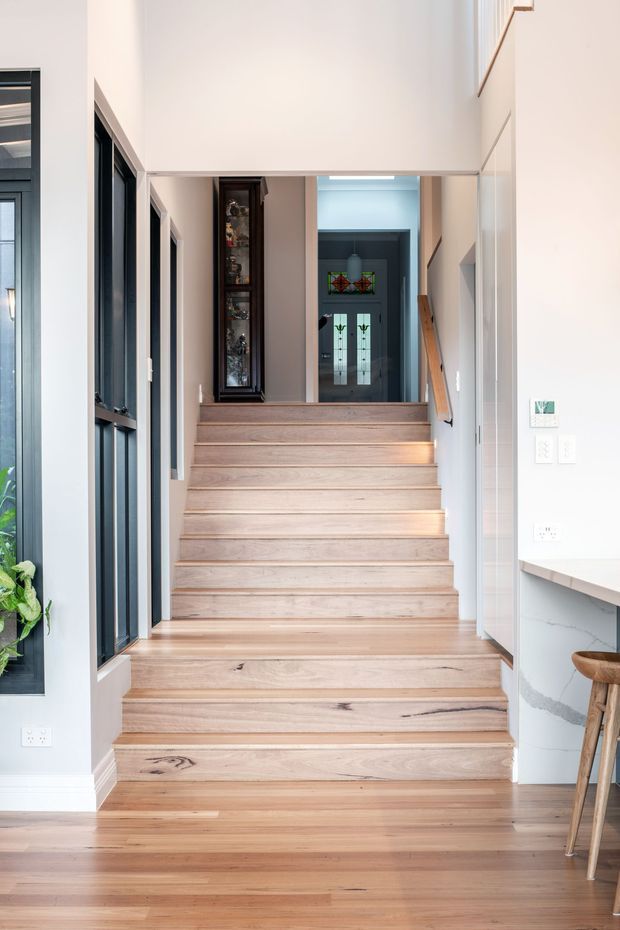 Bright interior view of a timber staircase rising through a hallway, with pale natural-wood treads and flooring, white walls, black-framed internal glazing, a slim timber handrail, and a dark stained built-in display cabinet. A kitchen counter and stool are visible at the right edge, while a glazed entry door is visible at the upper landing.