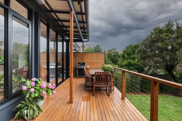 A timber outdoor deck and entertaining area beside a contemporary glazed home. The deck features warm timber boards, a timber balustrade with horizontal wire infill, a covered roof with exposed timber lining, outdoor dining furniture, a built-in barbecue area, and large black-framed glass doors opening to the interior. Lush planting and a residential garden sit beyond the deck beneath a dramatic overcast sky.