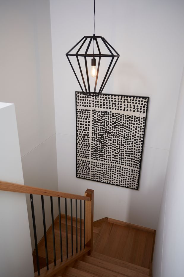 A stairwell with warm timber stairs and flooring, a timber handrail, and black vertical metal balusters. A geometric black pendant light hangs above a large monochrome dotted artwork mounted on a white wall. The space is clean and contemporary with a strong graphic accent.