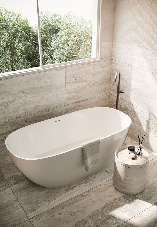 A bright, refined bathroom featuring a freestanding white bathtub, wall-mounted brushed metal tapware, large beige-grey stone-look wall and floor tiles, and a large window overlooking leafy greenery. A small cylindrical stone side table holds toiletries and a diffuser.