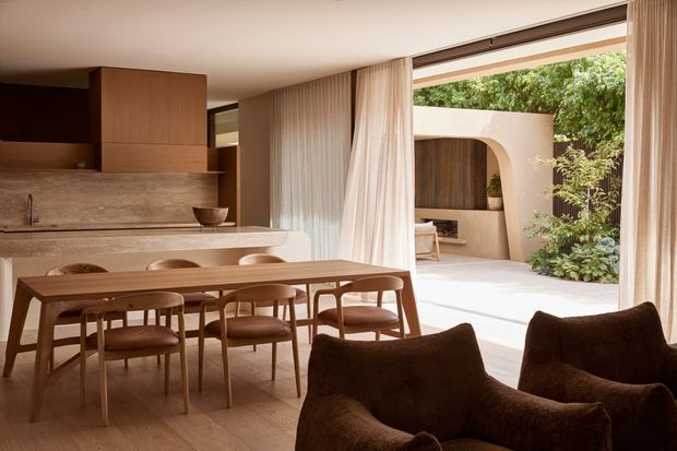 Warm contemporary open-plan kitchen and dining area opening onto a leafy courtyard. The room features a timber dining table with upholstered chairs, sculptural dark lounge chairs in the foreground, pale timber flooring, a stone kitchen benchtop and splashback, timber cabinetry, full-height sheer curtains, and large glazed openings. A curved plastered outdoor wall and lush planting are visible beyond.