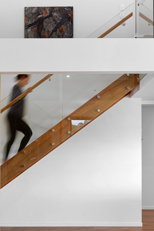 Interior view of a modern stairway with a broad white structural wall, warm timber stair stringer and handrail, and clear glass balustrades secured with exposed metal fittings. A blurred person is walking on the stairs, and abstract artwork is visible on the upper landing. Timber flooring and recessed ceiling lighting complete the clean, contemporary composition.