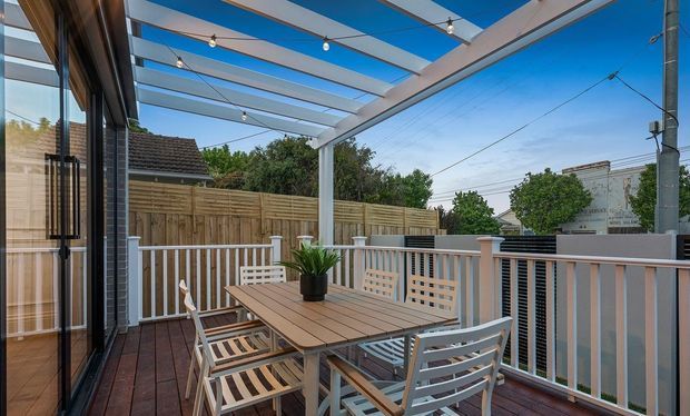 A sunny elevated outdoor dining deck beside a home, furnished with a rectangular timber dining table and slatted chairs. The deck has white-painted balustrades, a timber privacy fence, and a modern pergola with translucent roofing and festoon lighting. Neighbouring houses and greenery are visible beyond the railing.
