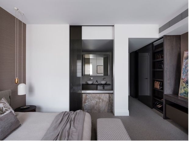 Contemporary bedroom with a partially visible upholstered bed, layered neutral bedding, pendant lighting, and a textured bench. A dark, finely detailed built-in wardrobe and shelving area lines the right side, while a compact ensuite vanity is visible through a black-framed opening at the centre. The room features crisp white walls, charcoal and timber-toned joinery, grey carpet, and a stone-clad vanity front.