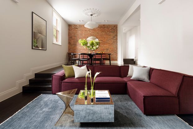 Bright open-plan living and dining interior with a large burgundy sectional sofa in the foreground, dark timber flooring and stairs, a grey area rug, and a metallic glass coffee table. The dining area sits against an exposed red-brick feature wall, with black dining furniture, a round wall mirror, pendant lighting, decorative ceiling detailing, and tall windows providing natural light.
