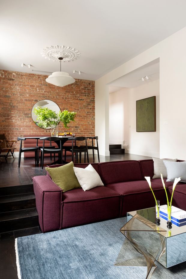A stylish open-plan living and dining area in a renovated interior. The foreground features a deep burgundy sectional sofa, pale blue-grey rug, mirrored geometric coffee table, and neutral cushions. A raised dining zone sits behind it with a dark dining table and chairs, a large round mirror, and leafy branches. The exposed red brick feature wall contrasts with white walls and ceiling, while a decorative ceiling medallion and contemporary pendant light add architectural character. Black timber flooring and steps connect the spaces.