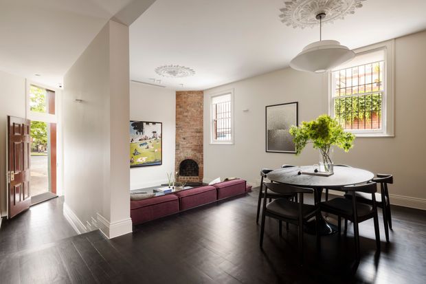 Bright, refined open-plan interior combining an entryway, living area and dining space. The room features dark timber flooring, pale painted walls, exposed brick around a fireplace, decorative ceiling medallions, tall leadlight-style windows, a round dining table with dark chairs, and a low burgundy sectional sofa. An open front door connects the interior to the exterior.