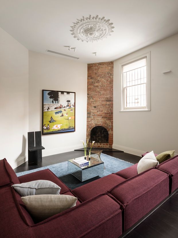 A refined living room with a large burgundy sectional sofa arranged around a square glass coffee table, set over a blue area rug. The room features dark timber flooring, pale walls, a white ceiling with an ornate decorative ceiling medallion, exposed red brickwork around a small arched fireplace, a tall barred window, framed artwork, and a sculptural black pedestal. The overall look combines historic architectural details with contemporary furnishings.