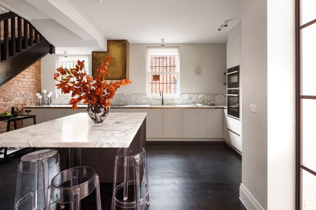Contemporary kitchen interior with a large marble island, dark timber cabinetry, pale cabinetry along the rear wall, integrated appliances, and transparent bar stools. A black-framed staircase and exposed brick wall add character, while a vase of orange foliage provides a warm accent. The room is bright with a restrained neutral palette and dark timber flooring.