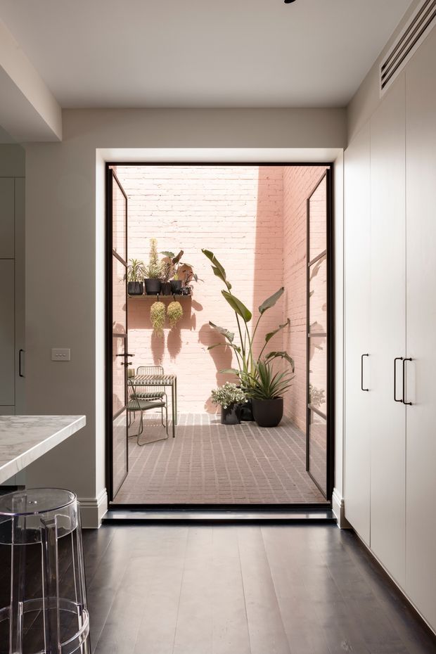 A refined contemporary kitchen interior opens through full-height glazed doors to a small enclosed brick courtyard. The kitchen features pale built-in cabinetry with dark handles, a light stone benchtop, dark timber-look flooring, and a clear acrylic bar stool. The courtyard has pale pink brick walls and matching brick paving, with potted tropical plants, wall-mounted planters, and a metal chair.