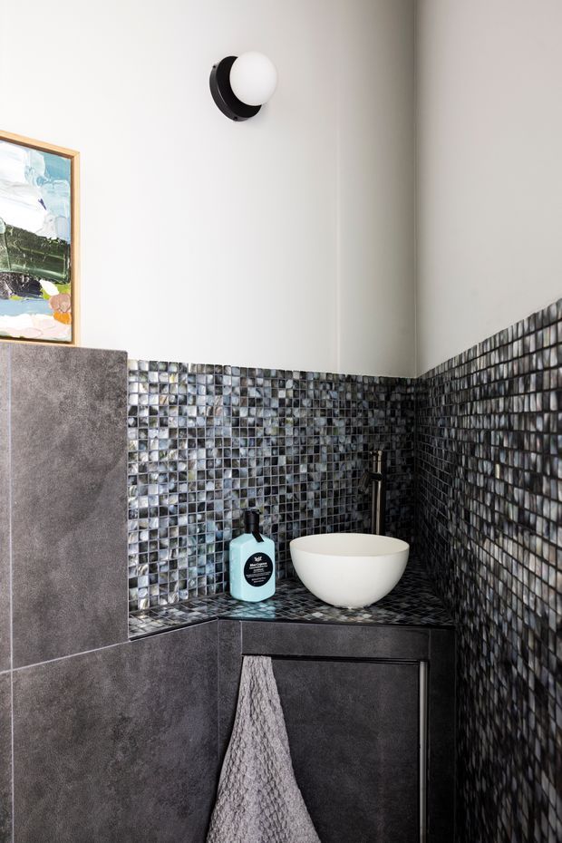 A compact contemporary bathroom vanity corner with dark grey stone-look wall and cabinet surfaces, a white vessel basin, black tapware, and glossy small-format mosaic wall tiles wrapping the corner. A round black-and-white wall sconce and framed artwork add decorative detail.