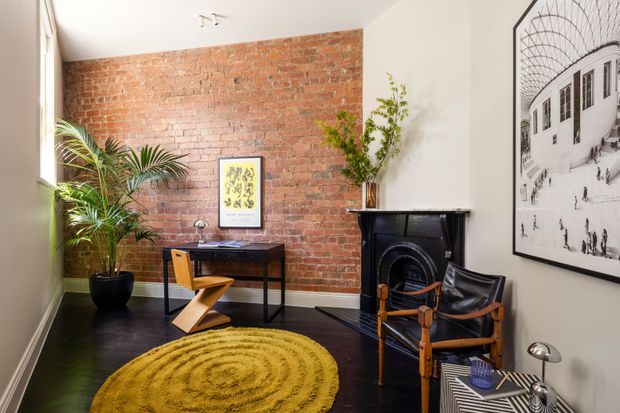 A compact, characterful study or living space with an exposed red brick feature wall, dark timber flooring, a black traditional fireplace, a slim desk, director-style chairs, indoor palms, and a large mustard circular rug. A black-and-white architectural artwork adds visual interest to the pale wall.