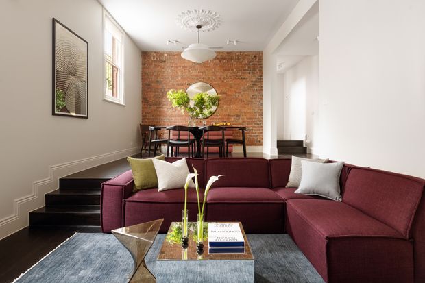 Bright, refined open-plan living and dining area with a deep burgundy sectional sofa in the foreground, a blue-grey rug, and a glass coffee table. A raised dining zone sits behind the living area, with a round table and chairs against an exposed red-brick feature wall. White walls, black stair treads, tall windows, contemporary lighting, and framed artwork complete the space.