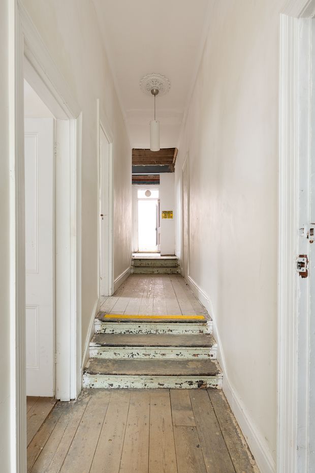 A narrow, bright residential hallway viewed from a lower landing, with worn painted timber floorboards, a short flight of distressed timber steps marked with yellow safety strips, white walls and traditional trim, multiple doorways, and a simple pendant light beneath an ornate ceiling rose. Exposed timber beams are visible near the far end, suggesting an older building under renovation.
