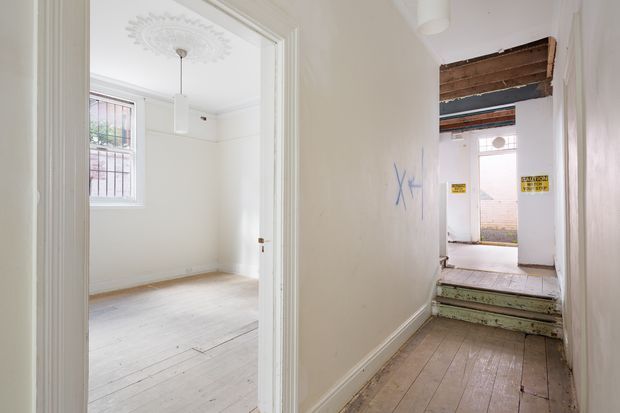 A narrow, worn hallway leads toward a rear entry or service area, with an open doorway revealing an empty bright room on the left. The space has painted white walls, aged timber floorboards, traditional trim and ceiling detailing, barred windows, and visible signs of renovation or disrepair, including exposed timber and warning notices near the far doorway.