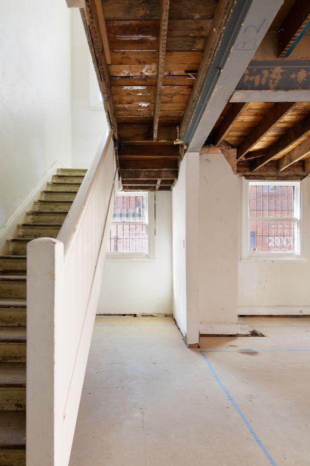 An unfinished interior under renovation, showing an exposed timber ceiling and structural beams, a partially stripped staircase, white plaster walls, tall multi-pane windows, and a bare concrete or unfinished subfloor marked with blue construction lines.