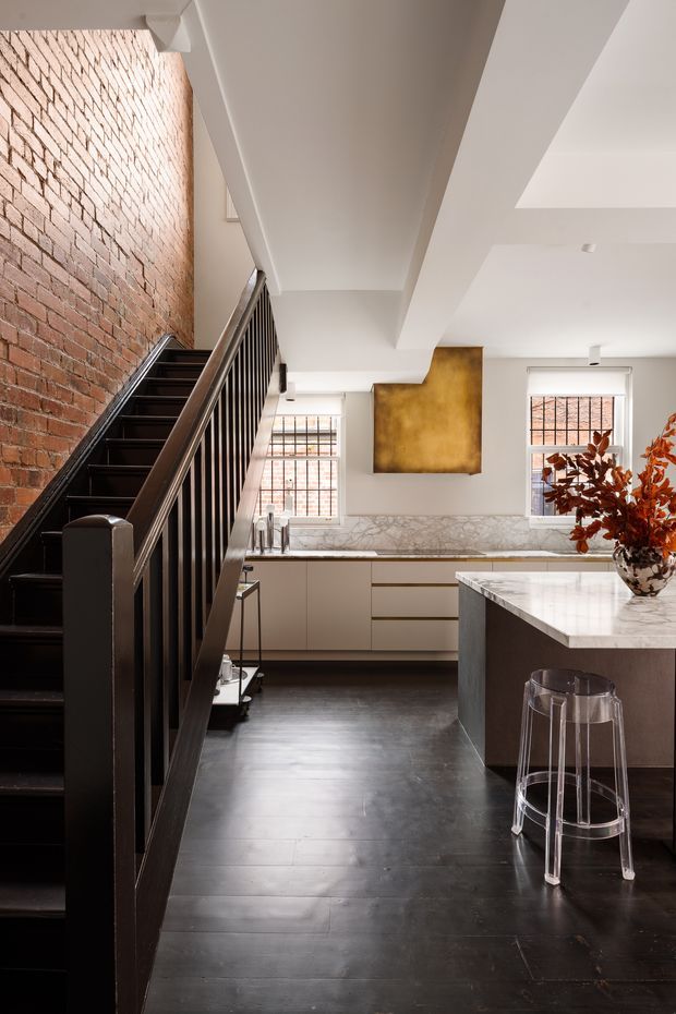 A refined kitchen viewed from beside a dark staircase. The space combines an exposed red-brick wall, dark timber flooring and staircase, pale built-in cabinetry, a marble-look benchtop and splashback, and a large island with a clear acrylic stool. White structural beams and ceiling planes create a bright, open interior, with brass-toned artwork and warm foliage adding contrast.