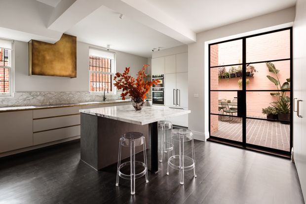Bright contemporary kitchen with a large marble island, integrated white cabinetry, dark timber flooring, black-framed glazing, and transparent bar stools. A black-framed glass door opens to a small brick-paved courtyard with planted greenery. A warm metallic feature panel adds contrast above the benchtop.