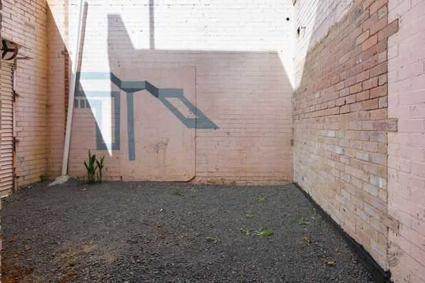 A small enclosed urban courtyard or service yard surrounded by pale, weathered painted brick walls. The ground is covered in dark gravel, with sparse weeds and a small plant at the edge. Strong sunlight casts angular architectural shadows across the rear wall, creating a stark, geometric scene with an industrial character.