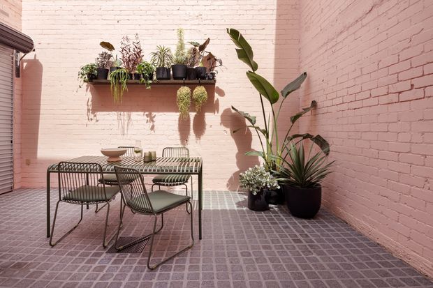 A compact enclosed courtyard or patio with pink-painted brick walls, a grey tiled floor, and abundant potted plants. A small black metal outdoor dining table with matching wire-frame chairs occupies the centre, while wall-mounted shelving displays additional plants. Strong sunlight creates crisp shadows, giving the space a warm, urban and botanical character.