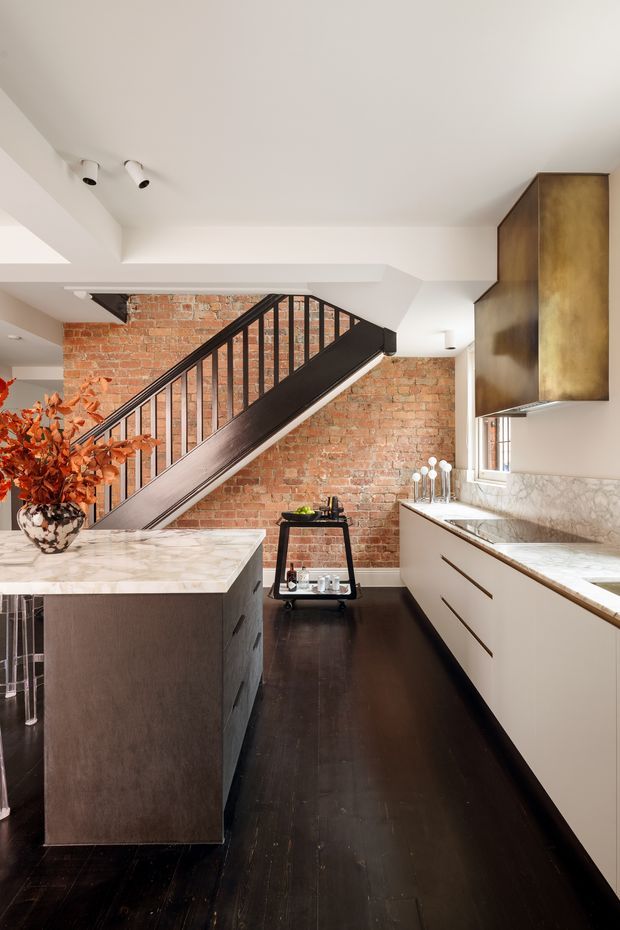 A sophisticated kitchen beneath an interior staircase, featuring a large marble island with dark cabinetry, matching marble benchtops and splashback, white lower cabinetry, dark timber flooring, exposed red brick walls, and a striking bronze-toned rangehood enclosure. A black metal stair balustrade and a small serving trolley add industrial character.