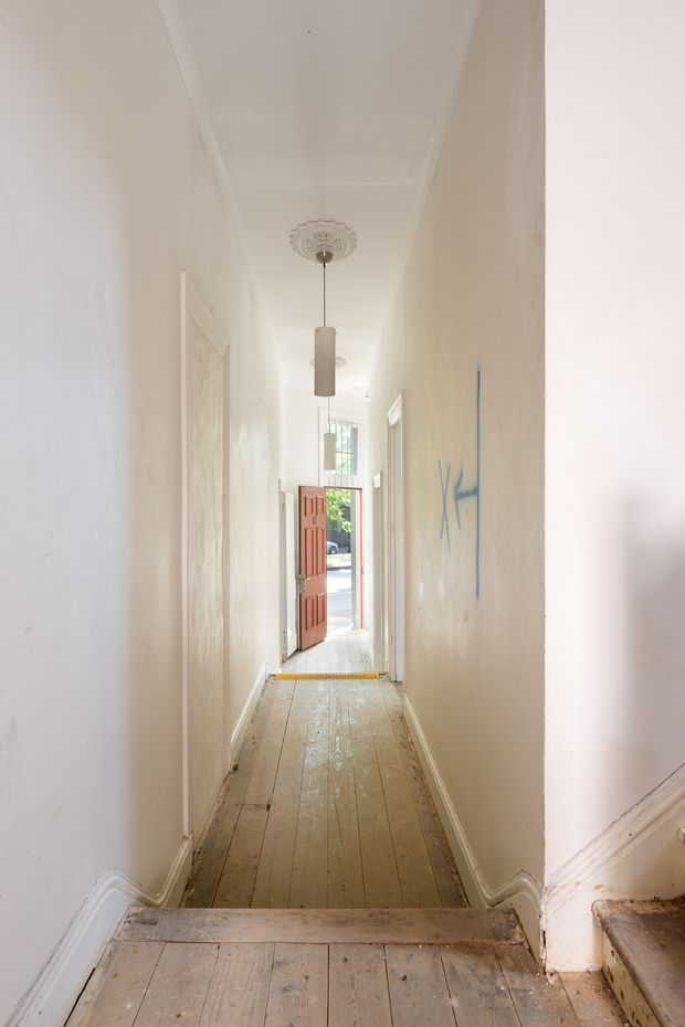 A narrow, elongated interior hallway leading to an open red-painted front door. The corridor has worn timber floorboards, white painted walls, decorative ceiling moulding with a central pendant light, traditional skirting boards, and a glimpse of the street outside. Blue graffiti is visible on the right wall.