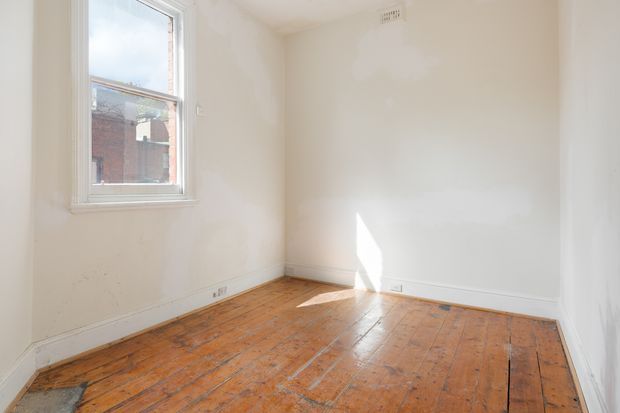 Bright, unfurnished small room with white painted walls and ceiling, a single white-framed window, white skirting boards, and an exposed warm-toned timber floor with visible plank seams and wear. Sunlight falls across the floor and wall, while a neighbouring brick building is visible through the window.