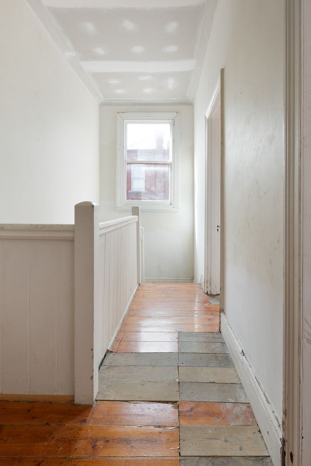 A narrow, bright upstairs hallway or landing with heavily worn painted timber floorboards, white painted walls and trim, a paneled stair balustrade, and a single white-framed window at the far end. Several doorways open from the corridor, and the ceiling has visible patching and recessed or reflected light spots. The space has a modest, aged residential character.