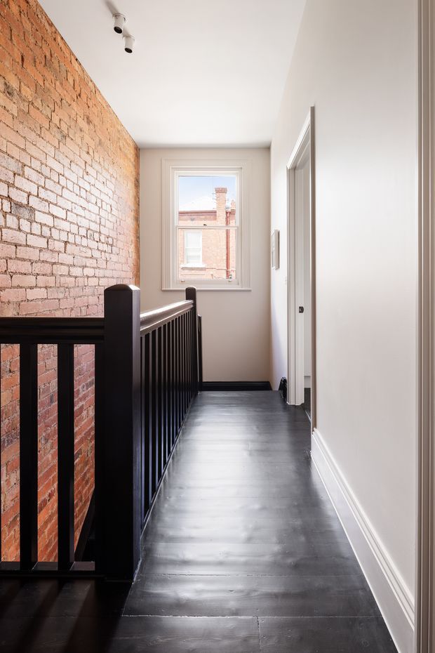 A narrow upper-level hallway or landing with dark painted timber flooring, white walls and trim, an exposed red brick feature wall, and a black timber stair balustrade. A window at the end provides natural light and views toward neighbouring brick buildings. The space has simple contemporary detailing with industrial character from the exposed masonry.