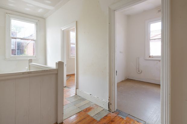 Bright, older residential interior showing a small hallway or landing with white-painted walls, trims and half-height balustrade. Open doorways lead to a sparsely finished room on the right and another room beyond, with original timber floorboards, a patch of multicoloured tiled flooring, timber-framed windows and simple traditional detailing.