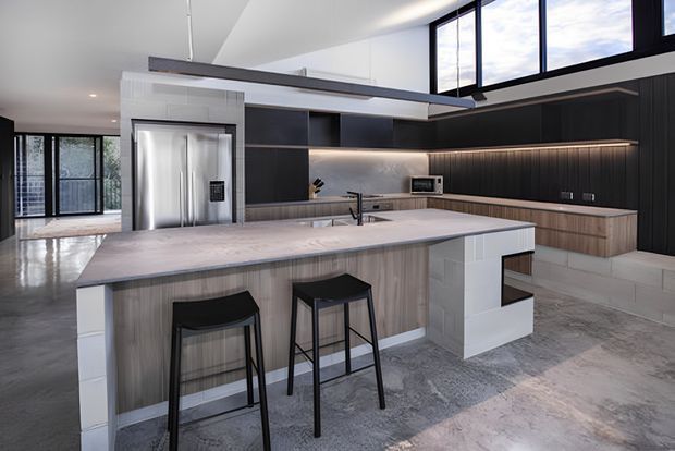 A spacious contemporary open-plan kitchen with a large island peninsula and two black bar stools. The kitchen features dark vertical-panel cabinetry, warm timber-toned lower cabinetry, a pale stone island benchtop, integrated stainless-steel appliances, under-cabinet lighting, high clerestory-style glazing, and polished concrete flooring. An adjacent open living or circulation area is visible beyond the kitchen.