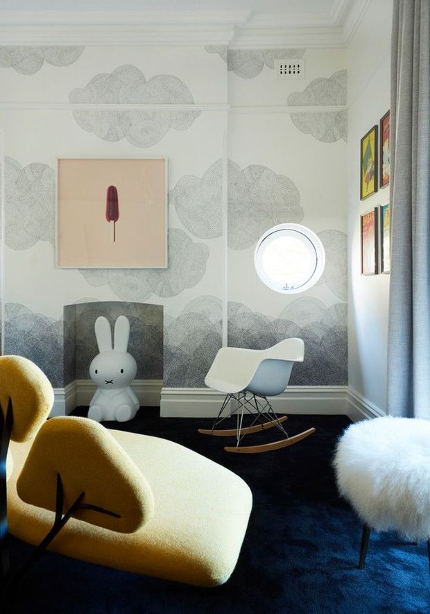 A playful, highly styled interior featuring a mustard-yellow sculptural lounge chair, a white furry stool, a white rocking chair, and a white rabbit figure arranged on a dark blue carpet. The room has ornate white ceiling mouldings, grey cloud-patterned wallcovering, framed colourful artwork, a round porthole-style window, and soft blue curtains. The overall aesthetic combines whimsical contemporary furnishings with classic architectural detailing.