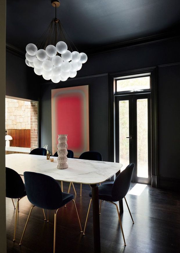 A dramatic, dark-toned dining room with black walls, ceiling and trim, dark timber flooring, and a black-framed glazed entry door. A large white marble dining table is surrounded by deep navy or black upholstered chairs with slender gold-toned legs. A sculptural cluster pendant of frosted glass globes hangs above the table, while a red abstract artwork and textured vase provide strong focal accents. A glimpse of a lighter adjoining space is visible through the opening at left.