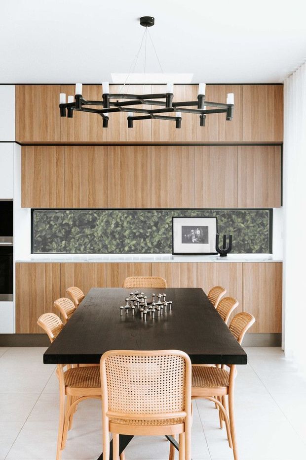 Bright contemporary dining area with a large dark rectangular dining table surrounded by woven cane chairs. Full-height warm timber cabinetry lines the rear wall, with a long dark botanical-patterned feature backsplash or artwork panel, white stone-like ledge, and a sculptural black multi-arm pendant light. White tiled flooring and white walls create a clean, high-contrast setting.