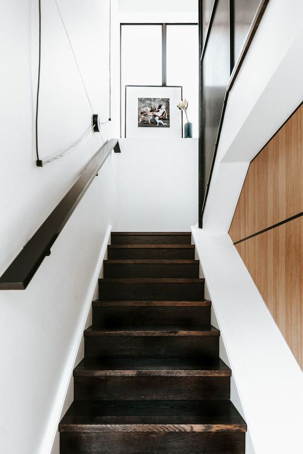 A narrow, modern interior stairway with dark timber treads, white walls, minimalist black handrails, and a timber-panelled wall on the right. The stairwell rises toward a bright landing with a framed artwork and simple decorative vase, with black-framed glazing and a dark mesh balustrade adding architectural detail.