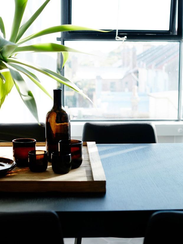 Bright, light-filled dining area with a dark rectangular dining table, black chairs, and a timber serving tray holding amber glassware and a large bottle. A leafy indoor plant sits beside large black-framed windows, creating a contemporary urban interior with strong natural light and high contrast.